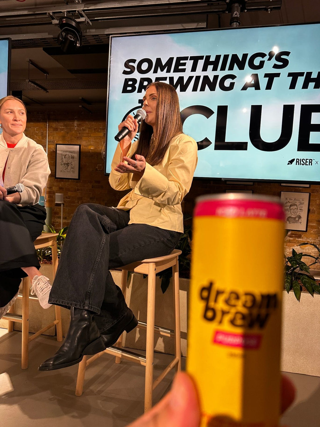 Aimee Squibb speaks at the 60-Second Club, with a DreamBrew can in the foreground and the RISER x DreamBrew event screen behi